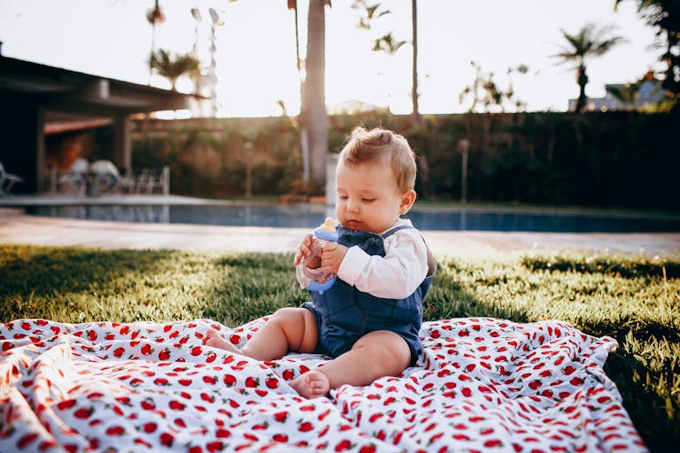 A cute baby sitting on a blanket in a sunny backyard, holding a bottle and enjoying a picnic.